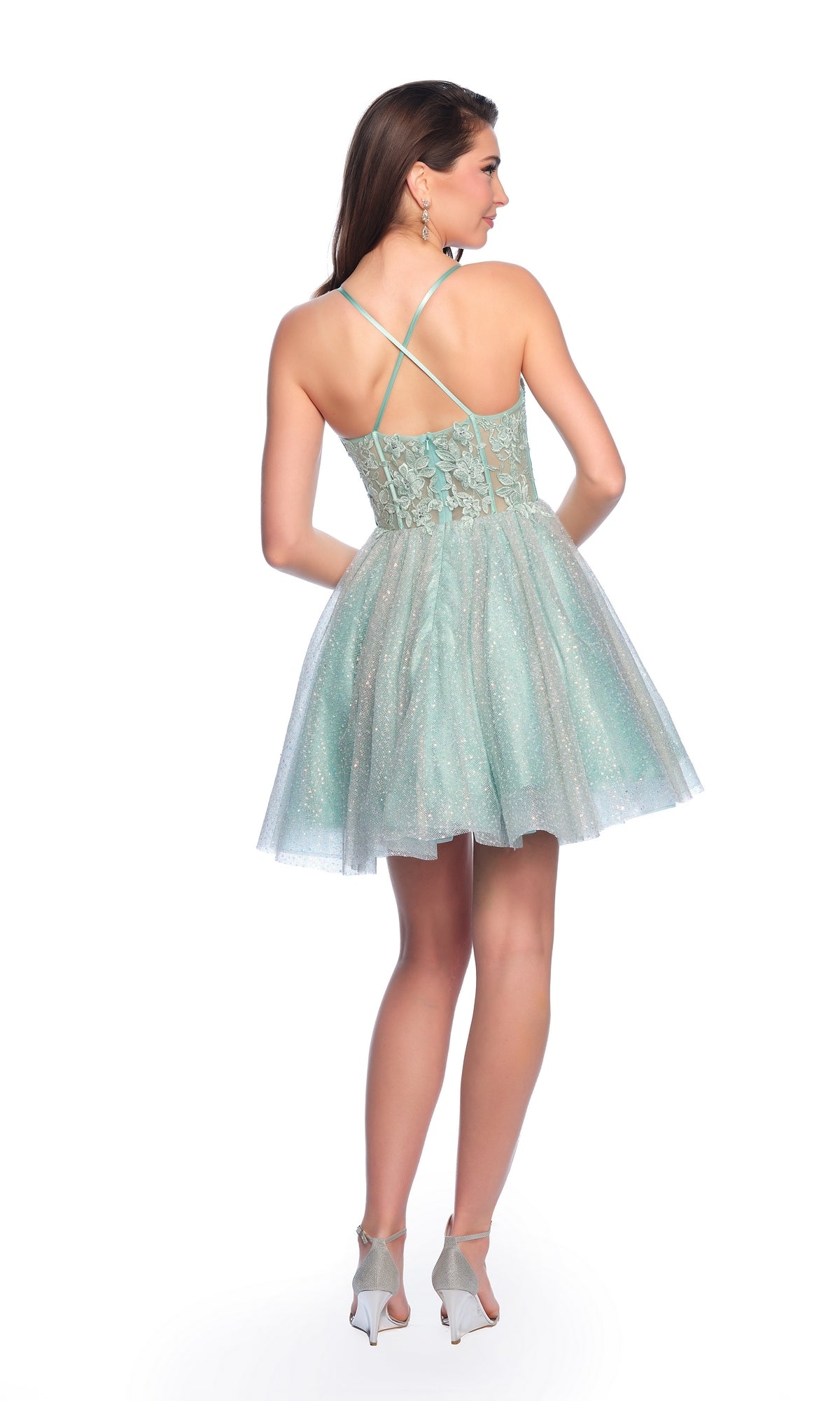 A woman stands with her back to the camera in a Dave & Johnny Short Glitter Hoco Dress 11808, featuring lace details and crisscross straps, paired with light heels against a white background.